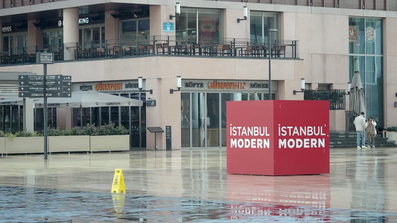 A modern urban plaza in Istanbul featuring the Istanbul Modern art museum signage and various commercial establishments, with wet ground reflecting the surroundings