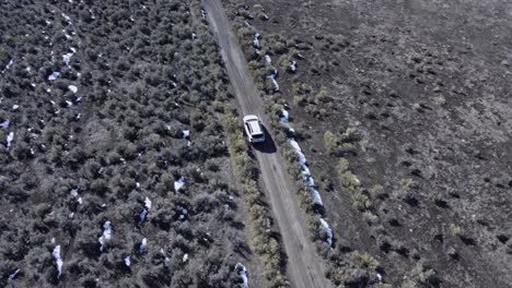 white suv offroading in the northern nevada desert on a sunny day in winter. aerial drone shot of the car driving through a shrub-covered hill, with a river to the left and a barren hill on the right.