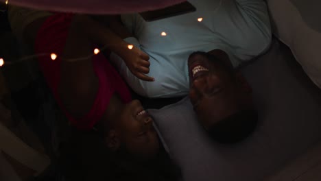 african american daughter and her father watching tablet lying in blanket tent with fairy lights