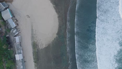 top down drone shot over tropical balangan beach peninsula in uluwatu bali indonesia with turquoise waves crashing over coral reef and rustic shacks lining beachfront