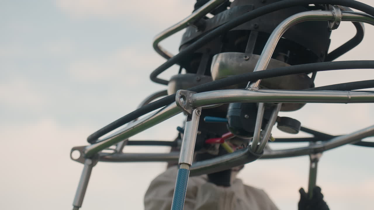 woman and assistant securing metal burner into vertical support poles of hot air balloon basket wearing gloves and protective jackets against red trailer backdrop set in grassy open field