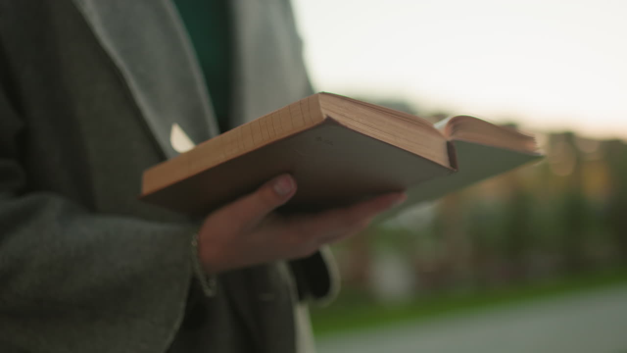 Close up of lady holding book with wrist chain visible, dressed in gray coat while reading outdoors, blurred background with wooden structure and greenery