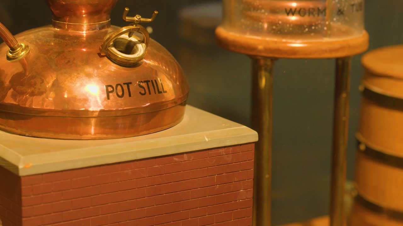Slow pan across copper pot still and worm tub distillation equipment under warm, indoor lighting