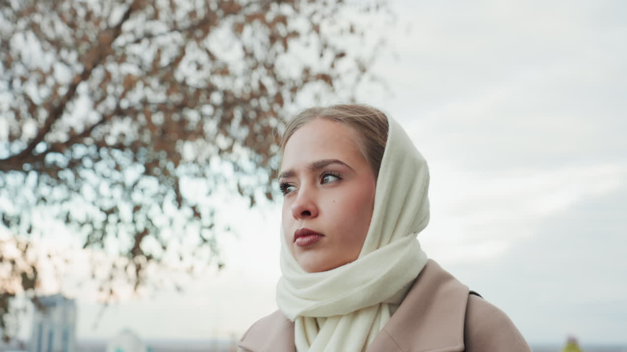 Mietiskelevä nainen ulkona. Tyyni nainen puiden ja kaupunkimaiseman keskellä. Mietteliäs nainen beigenvärisessä takissa kaupungin ja luonnon keskellä. Mietiskelevä nainen huivi kaulassaan puiden äärellä rauhallisessa kaupunkiympäristössä.