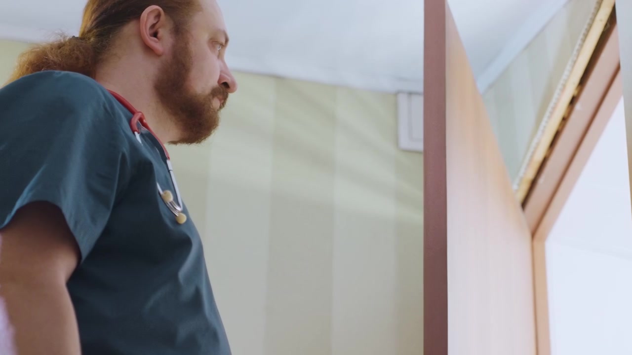 Paramedico bianco che entra nel corridoio di casa, medico dai capelli rossi in divisa verde acqua apre la porta con uno sguardo cauto, vista dal basso rivela interni e soffitto con carta da parati, atmosfera tranquilla e tesa durante un'emergenza