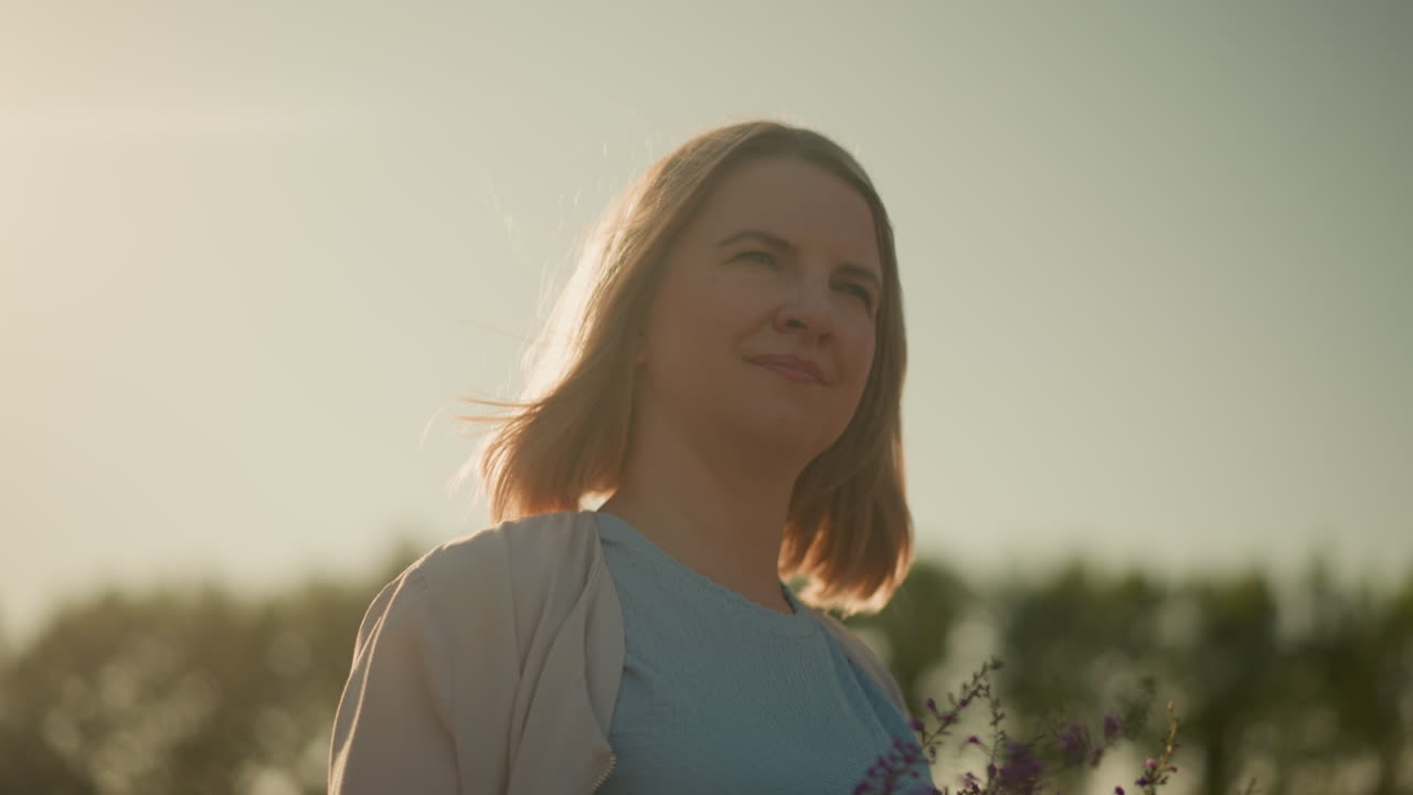 젊은 여성이 보라색 꽃을 들고 부드럽게 미소 짓고, 밝은 빛의 날, 빛이 그녀의 얼굴에서 반사되는 바에 서서 평화로운 순간을 즐기고 있습니다.