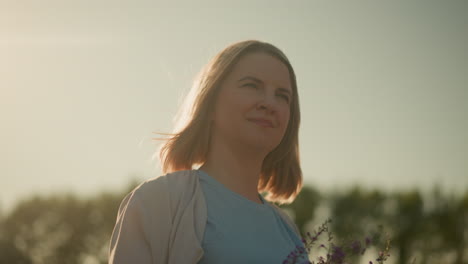 紫色の花束を手に持ってゆっくりと微笑んでいる若い女性のクローズアップ明るい晴れた日に屋外に立って太陽が彼女の顔に反射し平和な瞬間を楽しんでいます