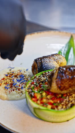 a dish of fried fish with tartar of fresh vegetables. cook's hand in black glove sprinkles the sauce with seeds and dried flower petals. close up. blurred backdrop. vertical video.