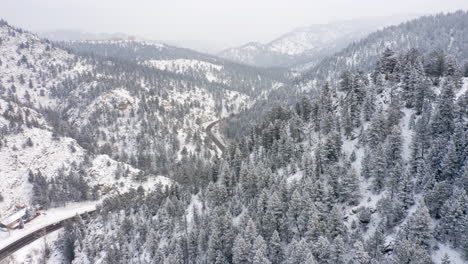Aerial-reveal-left-of-Boulder-Canyon-Drive-in-Colorado-during-the-winter-as-cars-drive-down-icy-road-surrounded-by-rocky-mountains-and-snow-covered-pine-trees