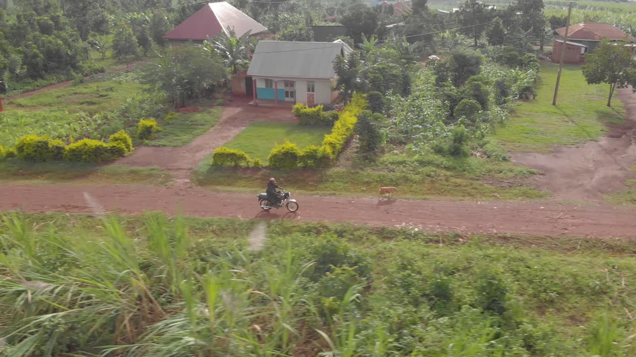 seguimiento de tomas aéreas desde el lado de un hombre africano con casco en una motocicleta que recorre un camino de tierra a través de un pueblo rural