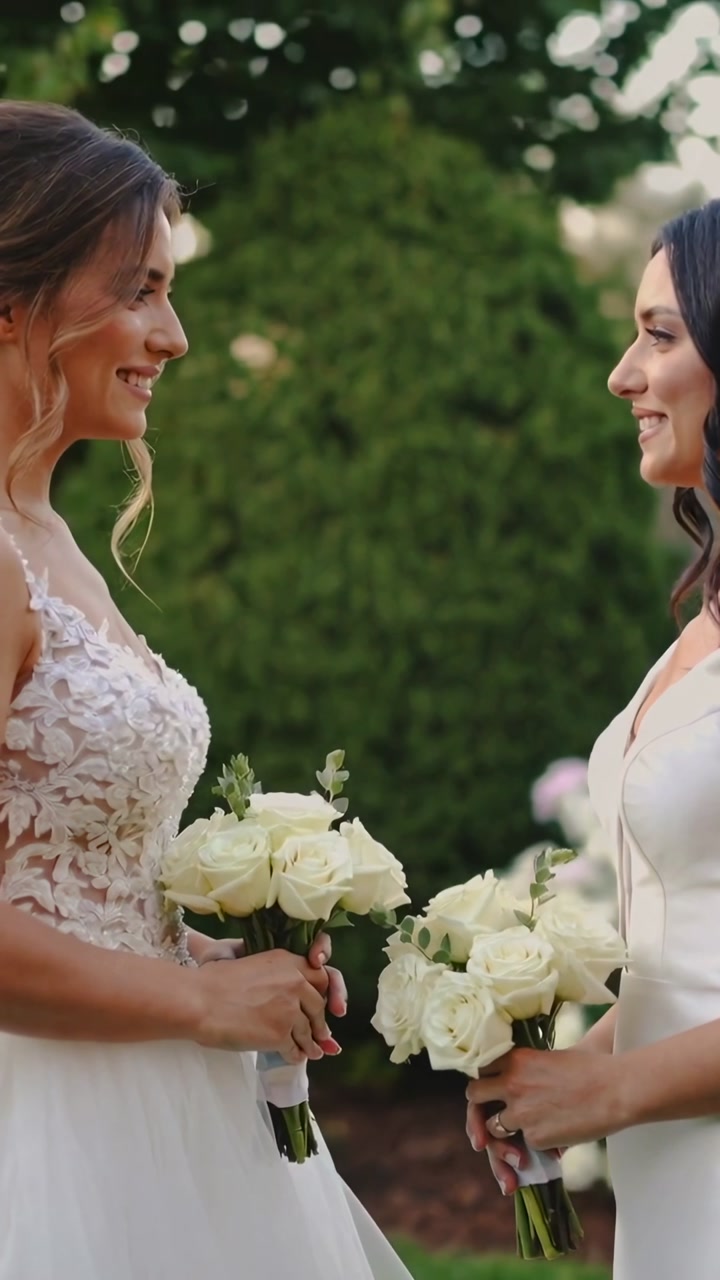 Vertical video: Exchanging smiles, two brides holding bouquets in garden, wearing gowns and rings