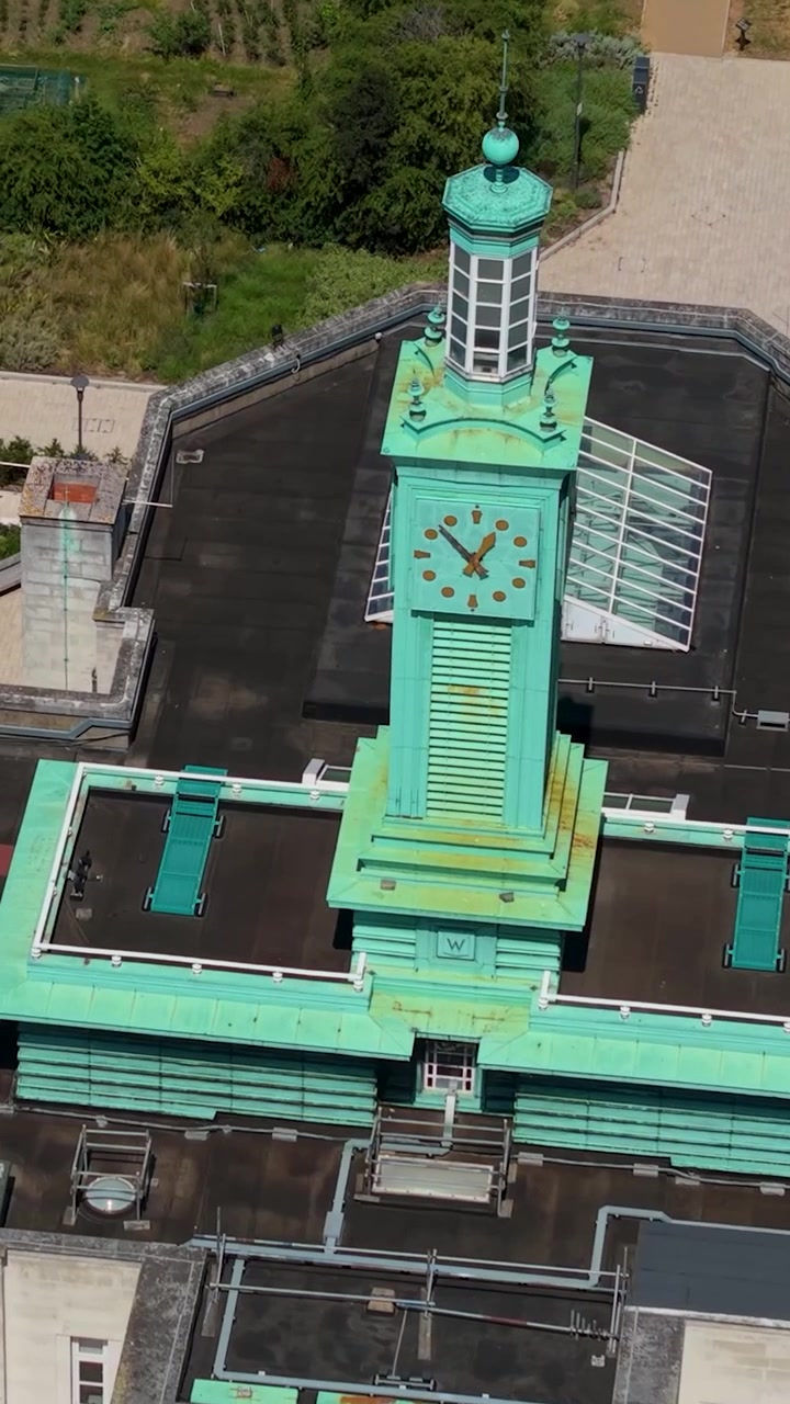 Green clock tower vertical drone orbit over Waltham Forest council offices, London. Aerial close rotation showing verdigris copper turret, rooftop solar panels, art deco architecture.