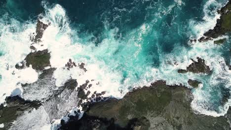 Luftaufnahme-Von-Meereswellen,-Die-Bei-Sonnigem-Wetter-Auf-Felsbrocken-Und-Korallenriffe-Am-Strand-Treffen,-Das-Wasser-Sieht-Blau-Aus-Und-Erzeugt-Weiße-Spritzer---Pengilon-Hill,-Indonesien,-Asien