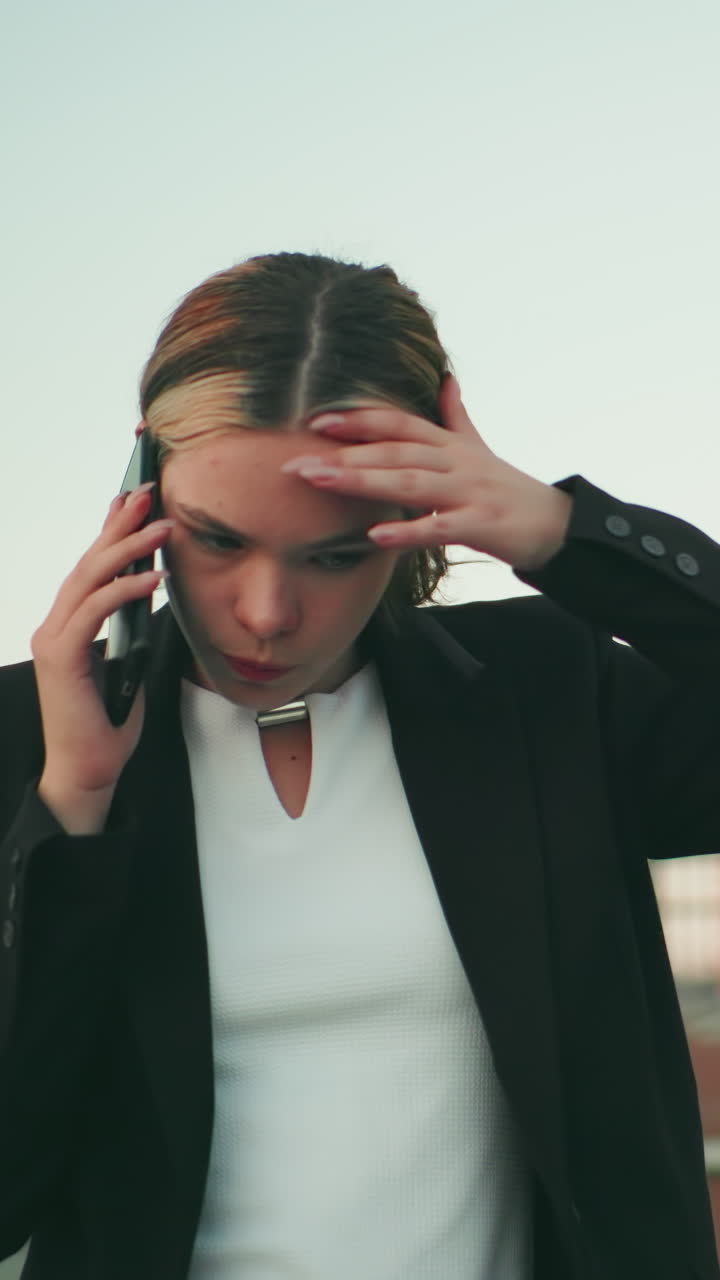 Tired white lady in professional outfit talking on phone with weary expression, walking near modern building, distant people blurred in background