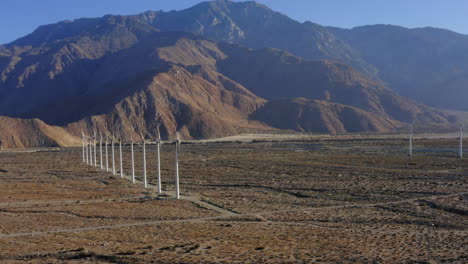 aerial view panning from right to left overlooking aligned wind turbines, desert and huge mountains near palm springs in the mojave desert, california, usa