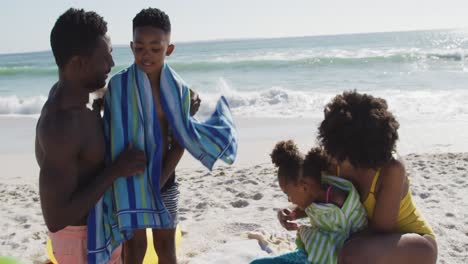 sonrientes padres afroamericanos quitando toallas de sus hijos en una playa soleada.