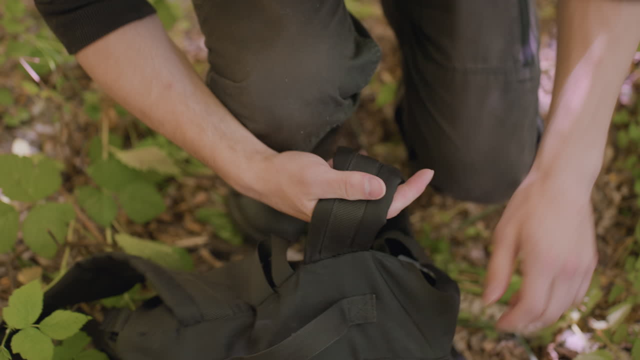Tourist secures bag while kneeling on forest ground, hands adjusting strap and zipper, phone tucked into dirty trouser pocket, leafy plants nearby, sunlight casting soft shadows on arm and pants