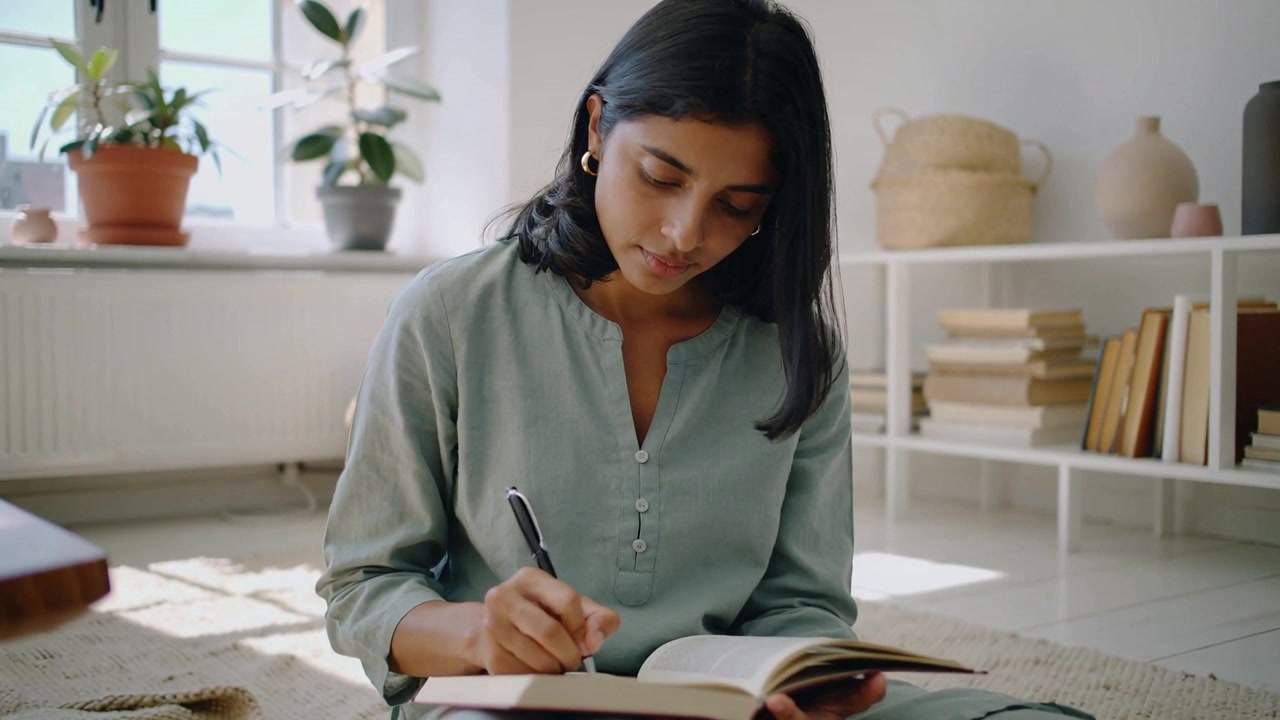 Writing woman in pale green top holding open book on lap near window, studying with pen