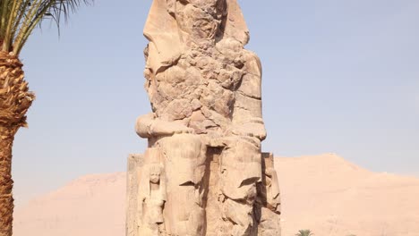 ruinas de la estatua gigante de faraón con montañas en el fondo en los colosos de memnon en luxor egipto con palma