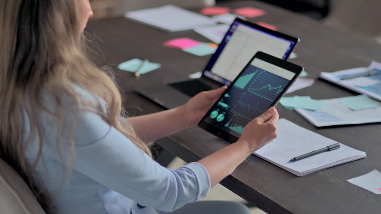 Woman in blue blazer analysing a graph on a tablet and writing in a notebook at a desk in an office