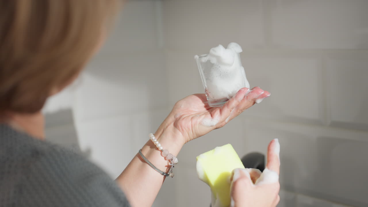 Rear view of woman pressing washing sponge foam inside glass cup while observing the cup in modern kitchen, with tile wall in background and soapy water in the glass