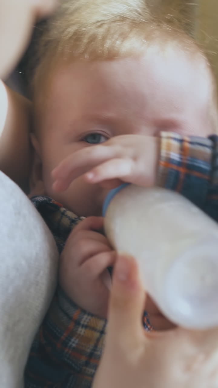 happy mommy rocks little son eating milk mix from plastic bottle with teat in arms against spacious light room close view