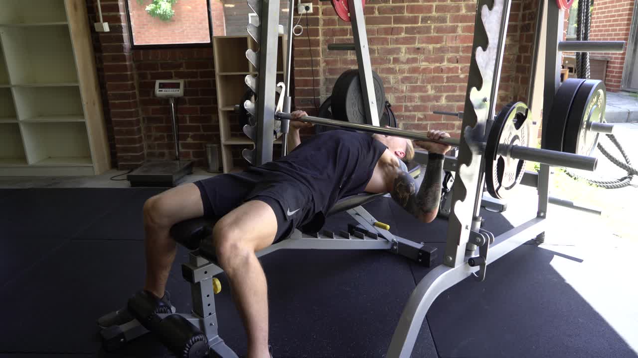 hombre tatuado en el gimnasio en casa máquina smith press de banca