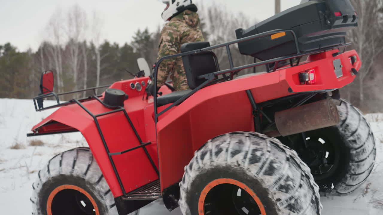 Pilota di ATV rosso, in un contesto invernale e innevato fuoristrada, con il quad rosso che naviga su un sentiero forestale innevato. Primi piani degli pneumatici mostrano il battistrada spesso intasato di neve. Il pilota in tenuta mimetica guida in salita attraverso un campo ghiacciato, sotto un cielo coperto.