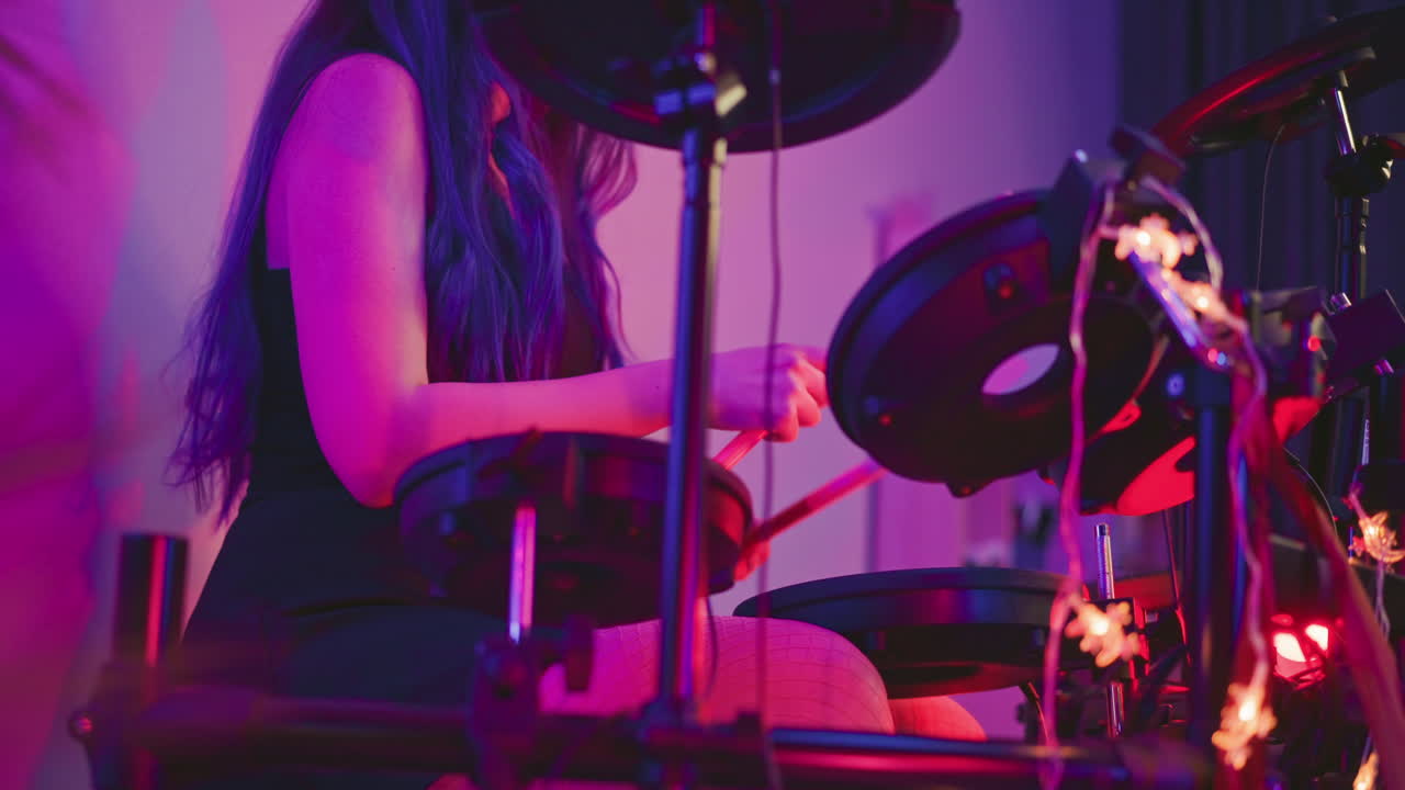 Lady playing electronic drum kit in warmly lit studio with vibrant purple and red glow, holding drumsticks while seated, surrounded by drum components and string lights
