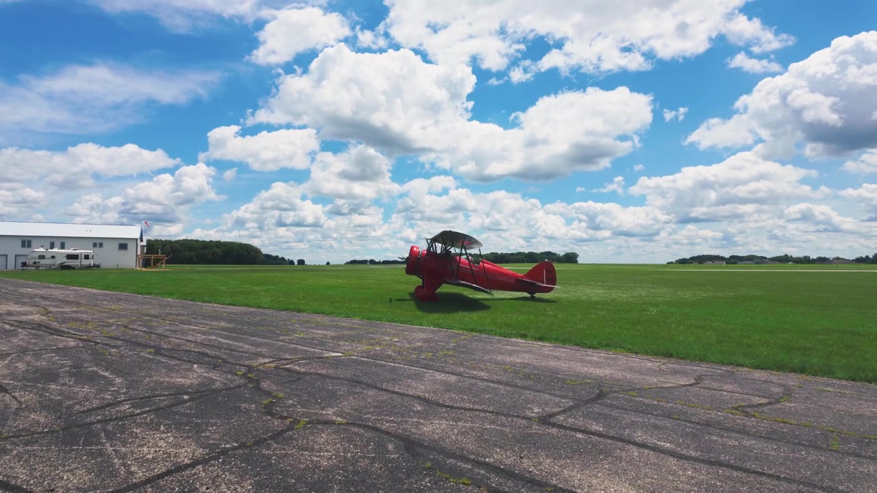 Un Waco rosso, aereo a elica, in rullaggio sull'erba sotto un cielo luminoso e nuvoloso.