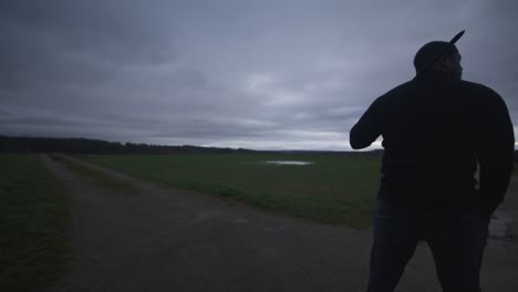 modern black swordsman warrior awaiting battle opponent in open field under grey sky, europe, 4k | muscular, intimidating, shadow, figure, blurry, dark clothes, powerful, hands in pocket, patrol