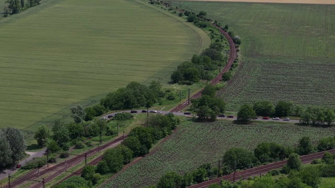 Una perspectiva aérea de dron captura una vía férrea cruzando una carretera rural entre extensos campos agrícolas cultivados en la Hungría rural