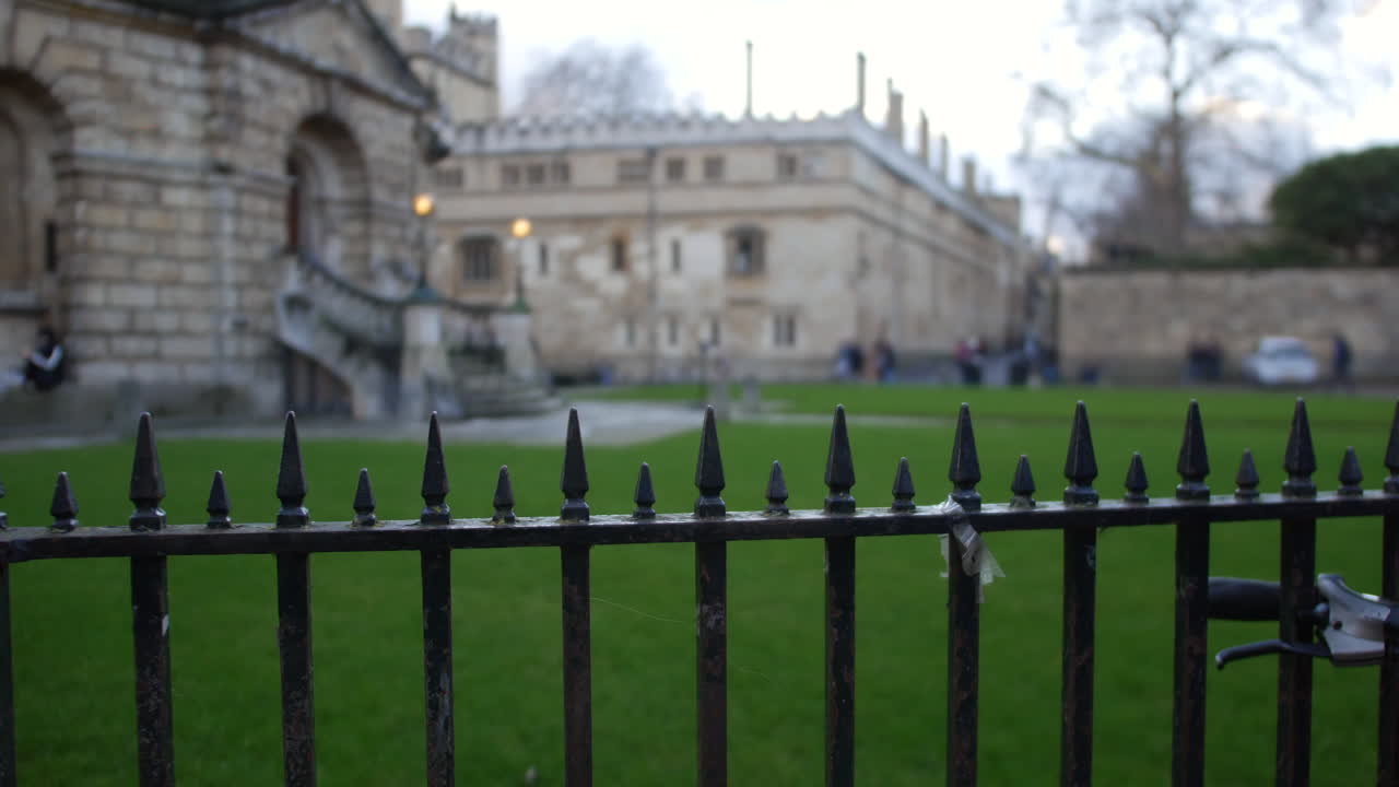 Ein verwitterter Eisenzaun umrahmt einen historischen Steingebäudekomplex in Oxford, England, ideal für touristische, akademische oder denkmalpflegerische Projekte, die europäische Architektur und ruhige Außenbereiche präsentieren.