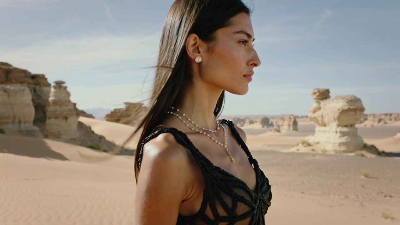 Brezza che solleva i capelli, modella che cammina indossando un abito nero intrecciato con tagli che mostra perle attraverso il deserto