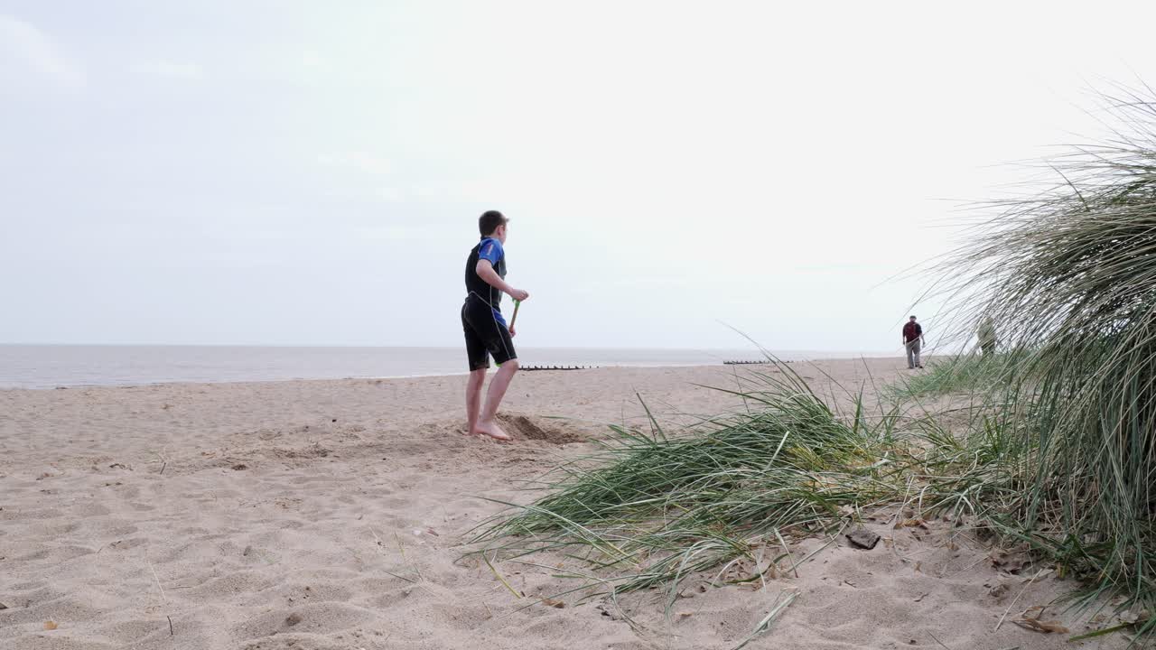 Young boy in a wetsuit on a beach digging in the sand. Playing with a surfboard and practicing skimming on the water. Seaside scene. Ocean waves breaking on the beach. Holiday time season