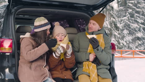 parents and daughter eating sandwiches and drinking hot drink sitting in the trunk of the car in a snowy forest