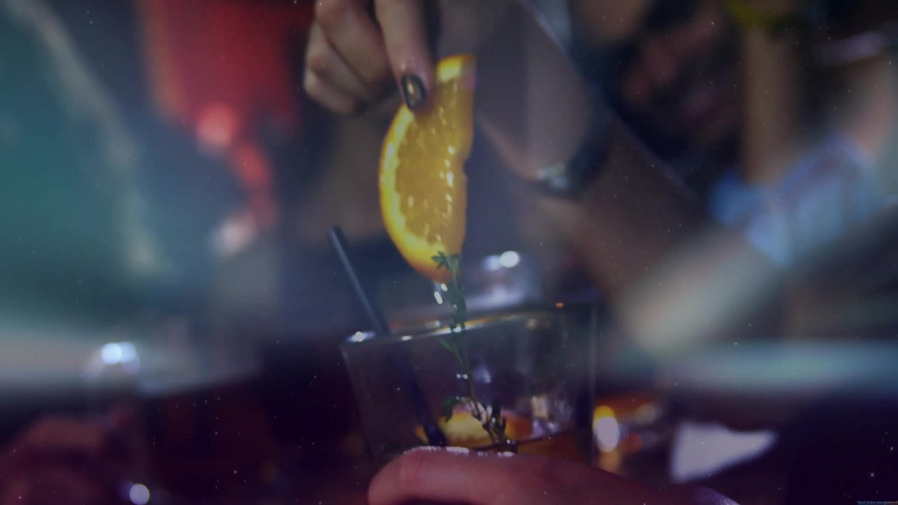 Mano di donna che si allunga per sistemare un'erba aromatica e posizionare una fetta d'arancia, mentre un uomo tiene un cocktail, preparazione al bar.
