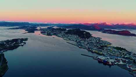 Toma-Aérea-De-La-Ciudad-De-Noruega-Aalesund-Durante-El-Crepúsculo-Con-Tono-Naranja-En-Las-Montañas-Cubiertas-De-Nieve