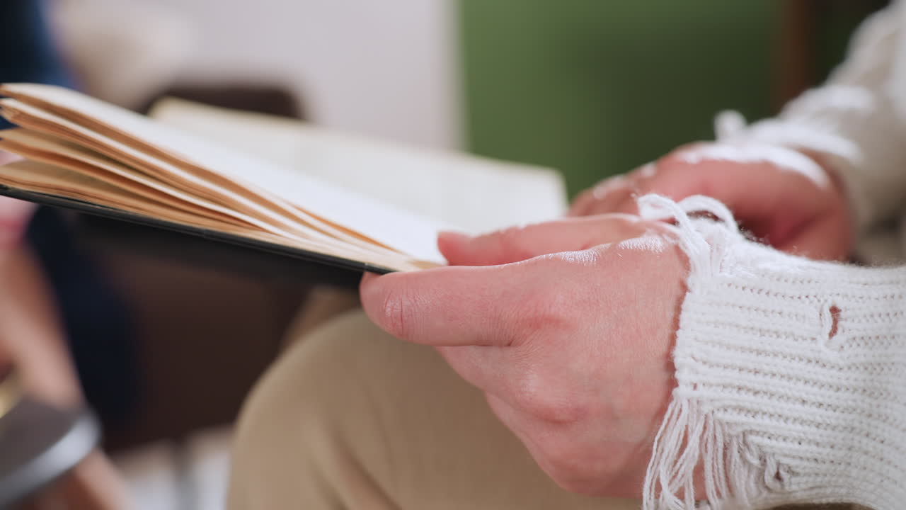 Close up of patient hands gently flipping through pages of old book while seated during calm therapy session, soft sweater sleeves visible, conveying reflection, healing, and emotional engagement