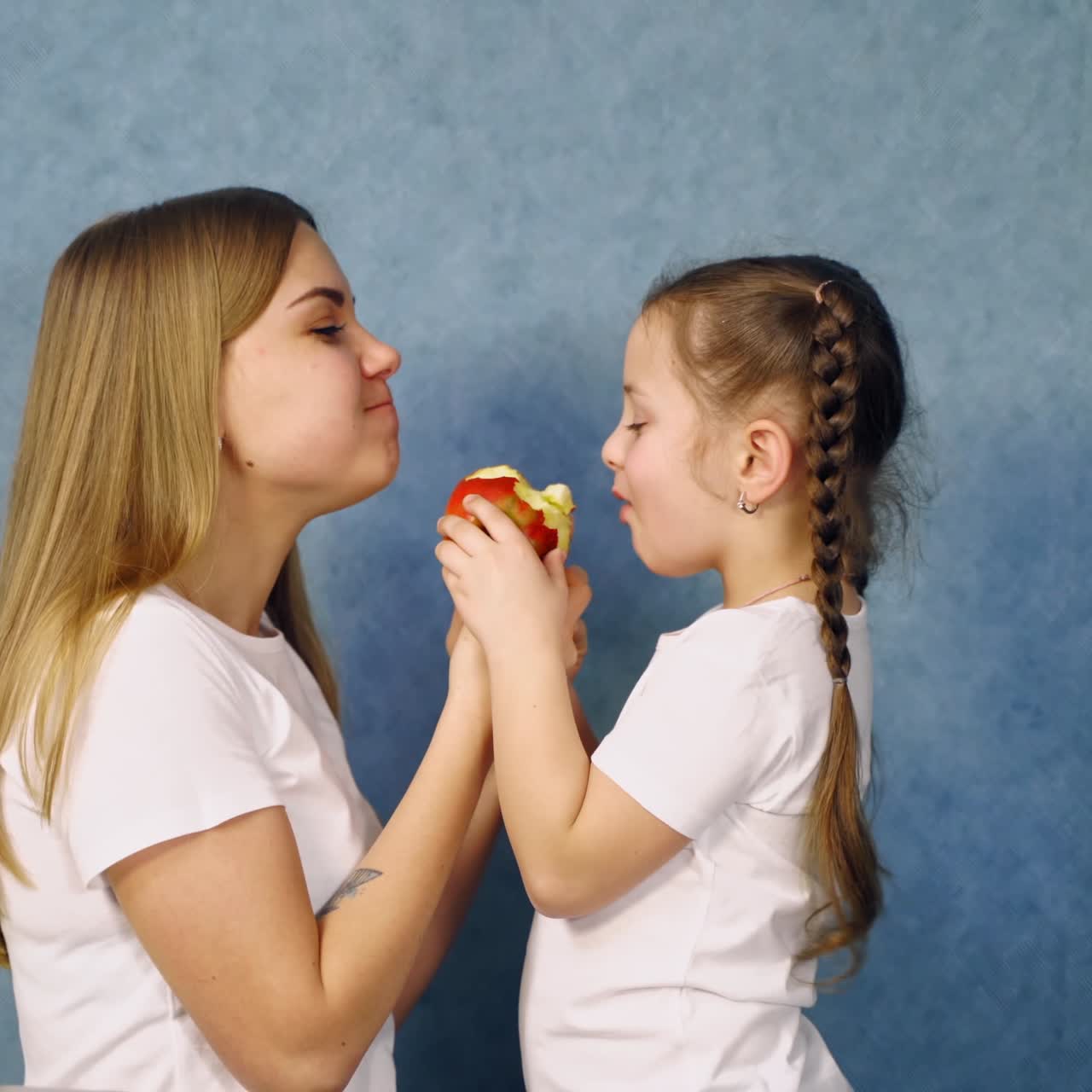 Little girl and her mother in white t-shirts bite an apple. Side view of mother and daughter eating one ripe apple on blue studio background.