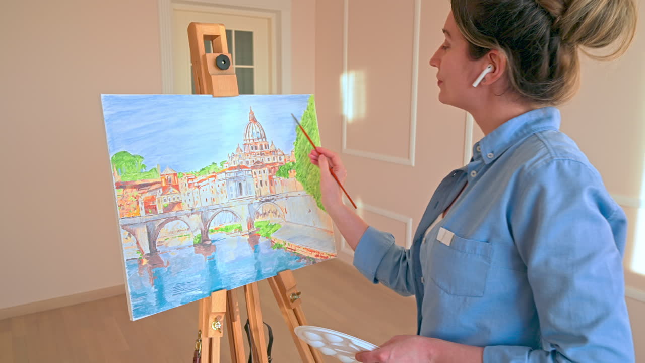 Caucasian woman painting a picture with a palette in her hands and listening music with earphones indoors