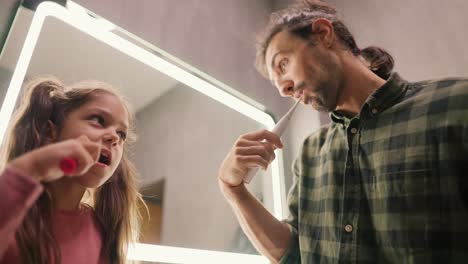 A-little-brunette-girl-with-a-ponytail-and-a-pink-dress-brushes-her-teeth-with-a-toothpick-along-with-her-single-father,-a-brunette-man-there-in-a-Green-checkered-shirt-near-a-mirror-in-a-modern-bathroom