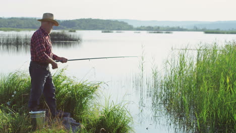 side view of a old man in a hat standing on the lake shore and fishing with a rod