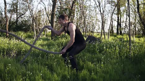 side view of an athletic male working out using battle ropes. crossfit. swing. green park. black sport clothes