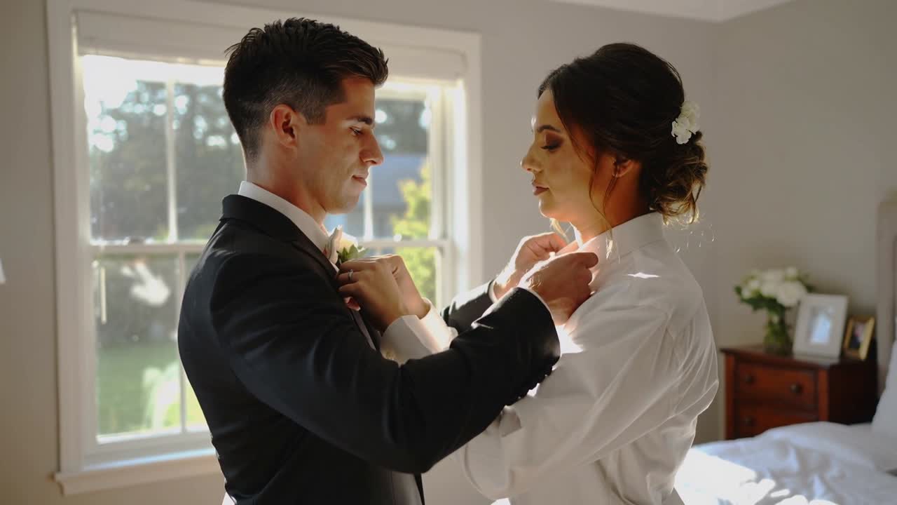 El novio asegurando un prendedor floral mientras la novia, ya vestida, levanta las manos y ajusta su cuello junto a la ventana antes de la boda.