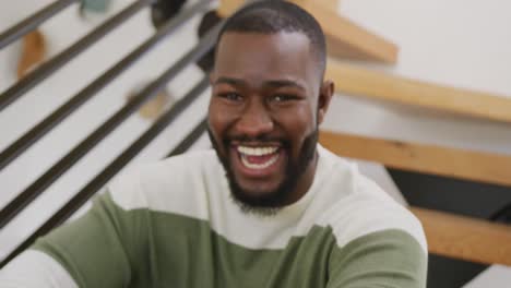 video portrait of happy african american man laughing to camera, sitting on stairs at home