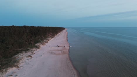 Car-Stuck-in-Sand-at-the-Dune-Sea-Coast-of-Baltic-Sea-Bernati,-Latvia-Sun-Going-Beyond-the-Horizon-Illuminating-Water-and-Clouds-in-the-Sky,-Calm-Sea-Waves,-Romantic-Mood,-Wide-Angle-Drone-Shot