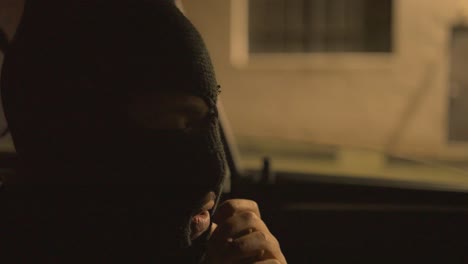 a black man sitting in the drivers seat of a car of a car adjusting a black ski mask over his face in front of a warehouse