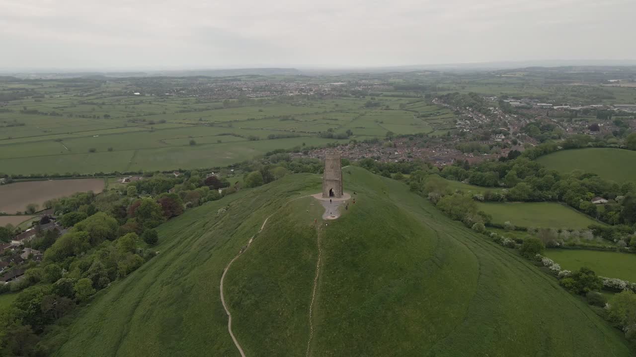 Aerial view of Glastonbury, drone moving backwards moving over and away from Glastonbury Tor, drone flying over the green fields, some houses on the left side of the camera, town in the background.