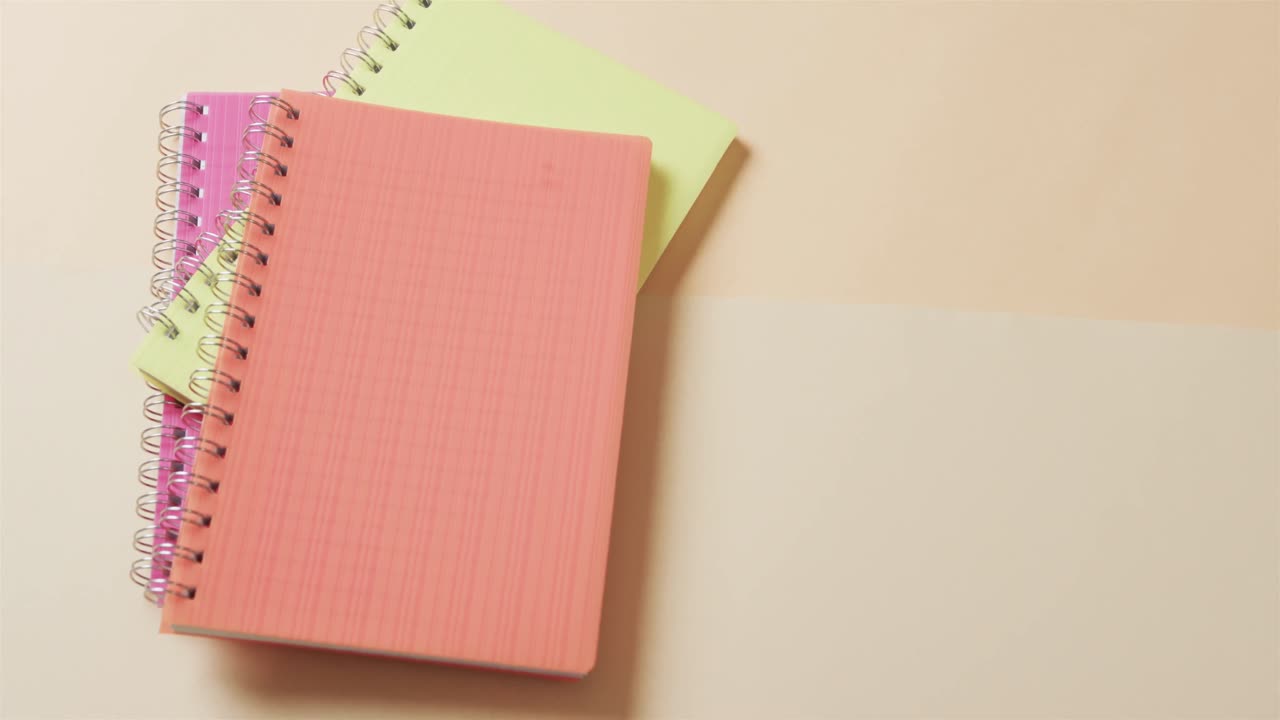 Overhead view of colourful notebooks with copy space on beige background, in slow motion